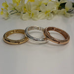 Three gold, silver, and rose gold bracelets on a white surface with yellow flowers in the background.