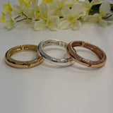 Three gold, silver, and rose gold bracelets on a white surface with yellow flowers in the background.