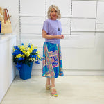 Woman wearing a purple top and colorful skirt standing next to a blue vase with flowers against a white wall.