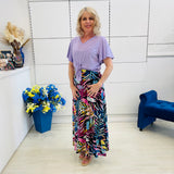 Woman in a light purple top and colorful skirt standing in a room with a blue chair and floral arrangement.