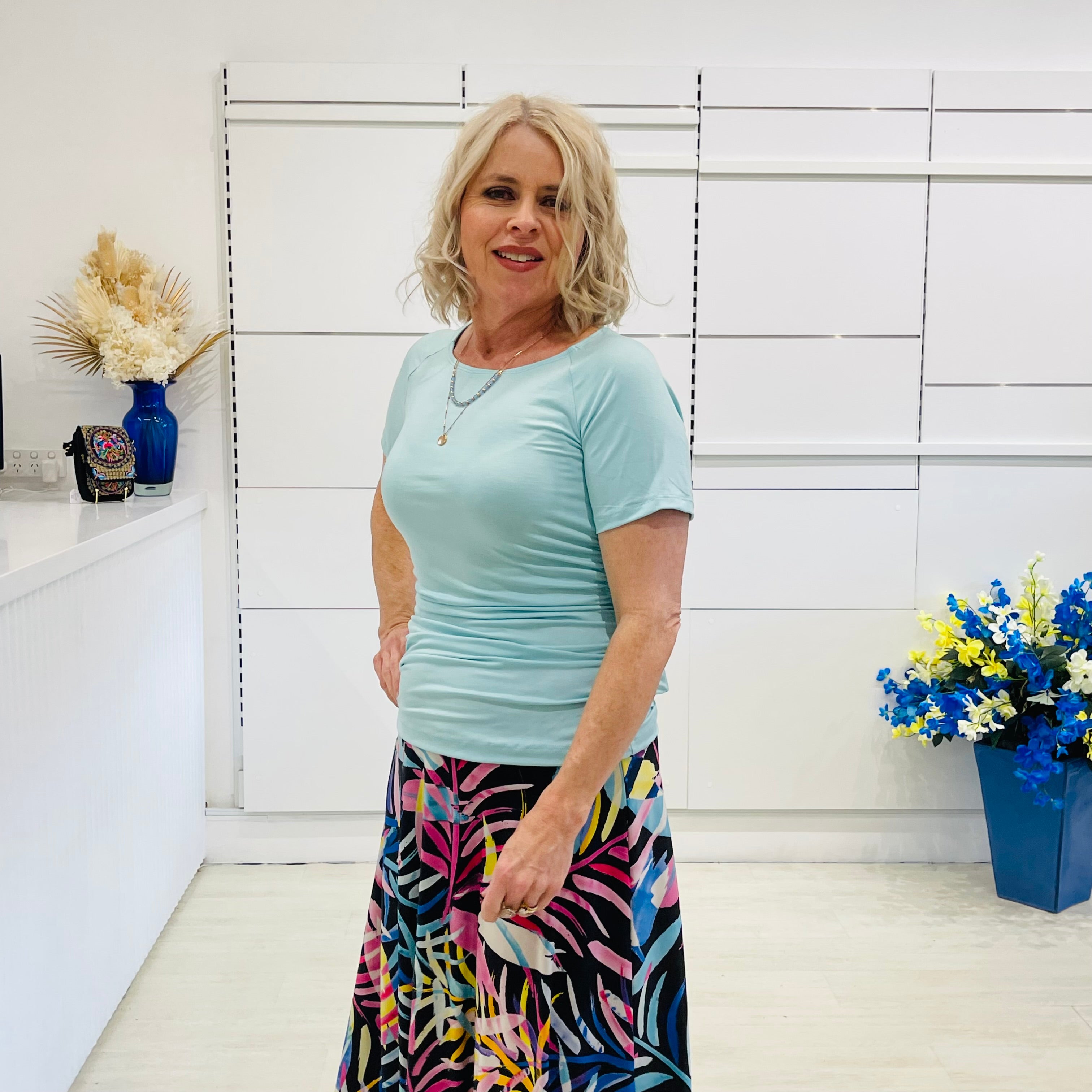 Woman in a light green  top and colorful skirt standing in a room with white walls and decorative elements.