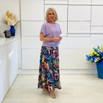 Woman wearing a light purple top and colorful skirt standing in a room with white walls and a vase of flowers.