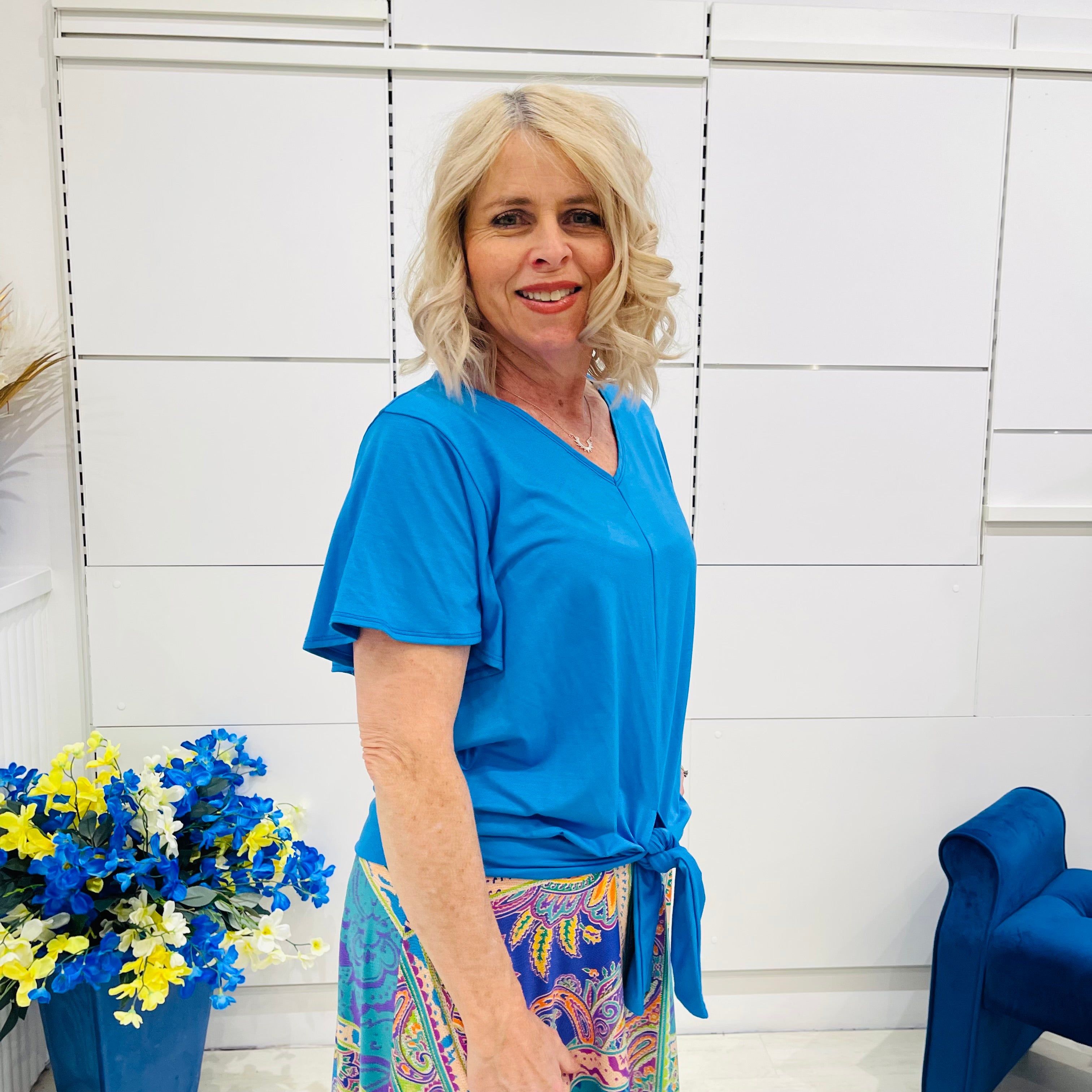 Woman in a blue top and colorful skirt standing next to a blue chair and floral arrangement against a white wall.
