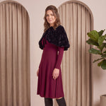 Woman in a burgundy dress with a black fur shrug standing in front of beige curtains.