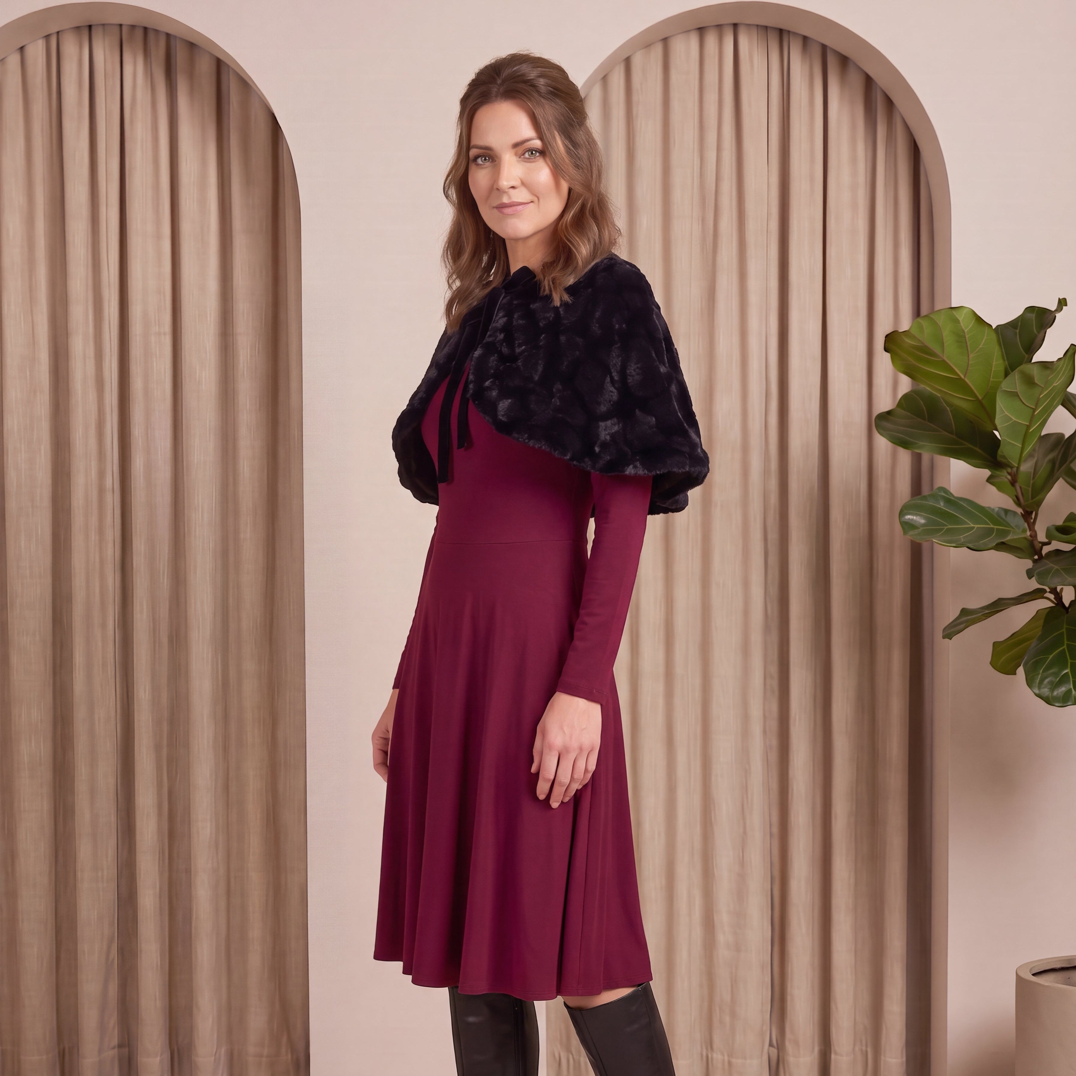 Woman in a burgundy dress with a black fur shrug standing in front of beige curtains.