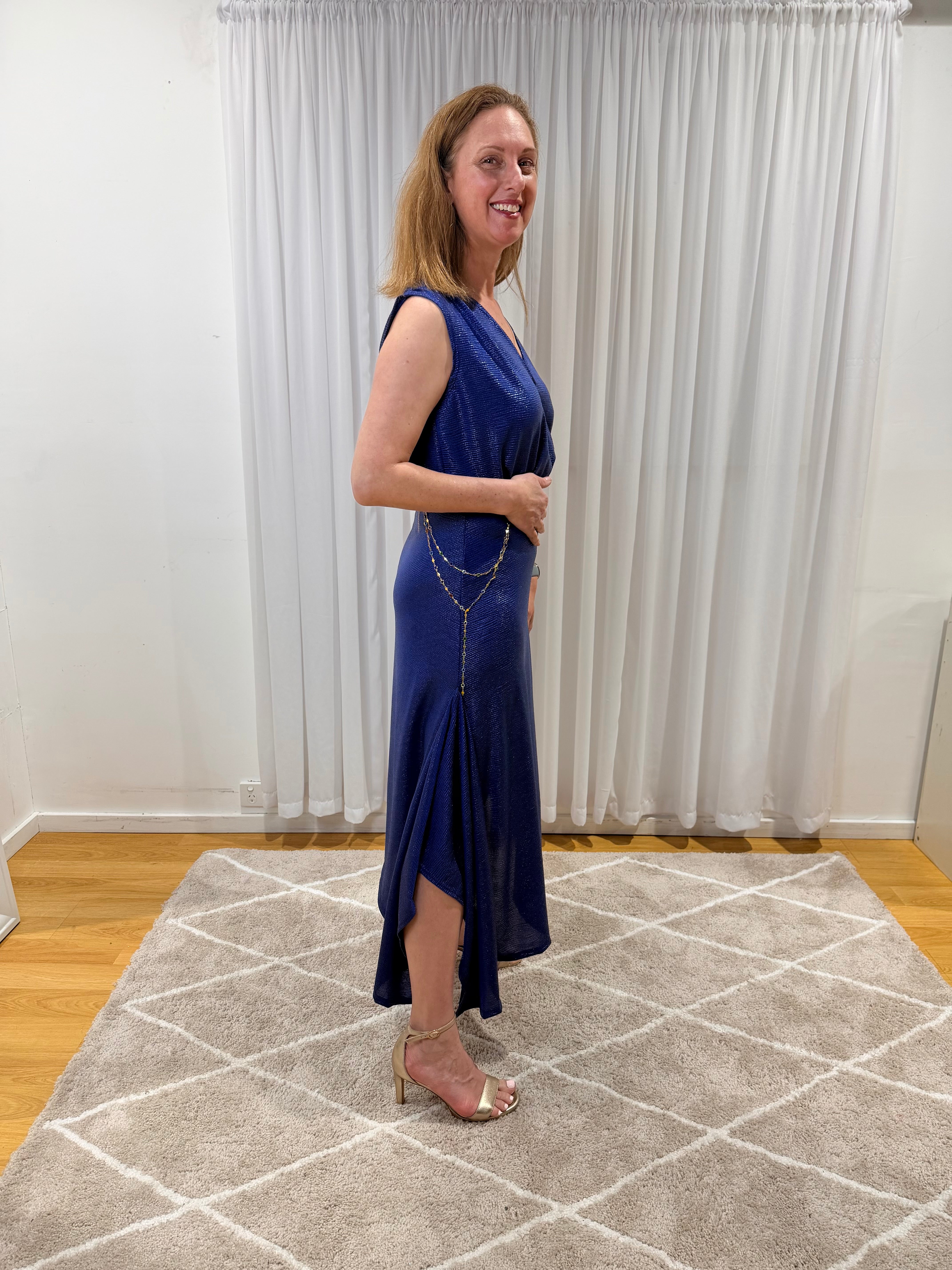 A woman modeling a sleeveless royal blue maxi dress with a V-neckline and a draped side slit, standing on a patterned rug in front of white curtains.