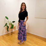 Woman wearing a black top and colorful patterned pants standing in a room with a plant and small table.