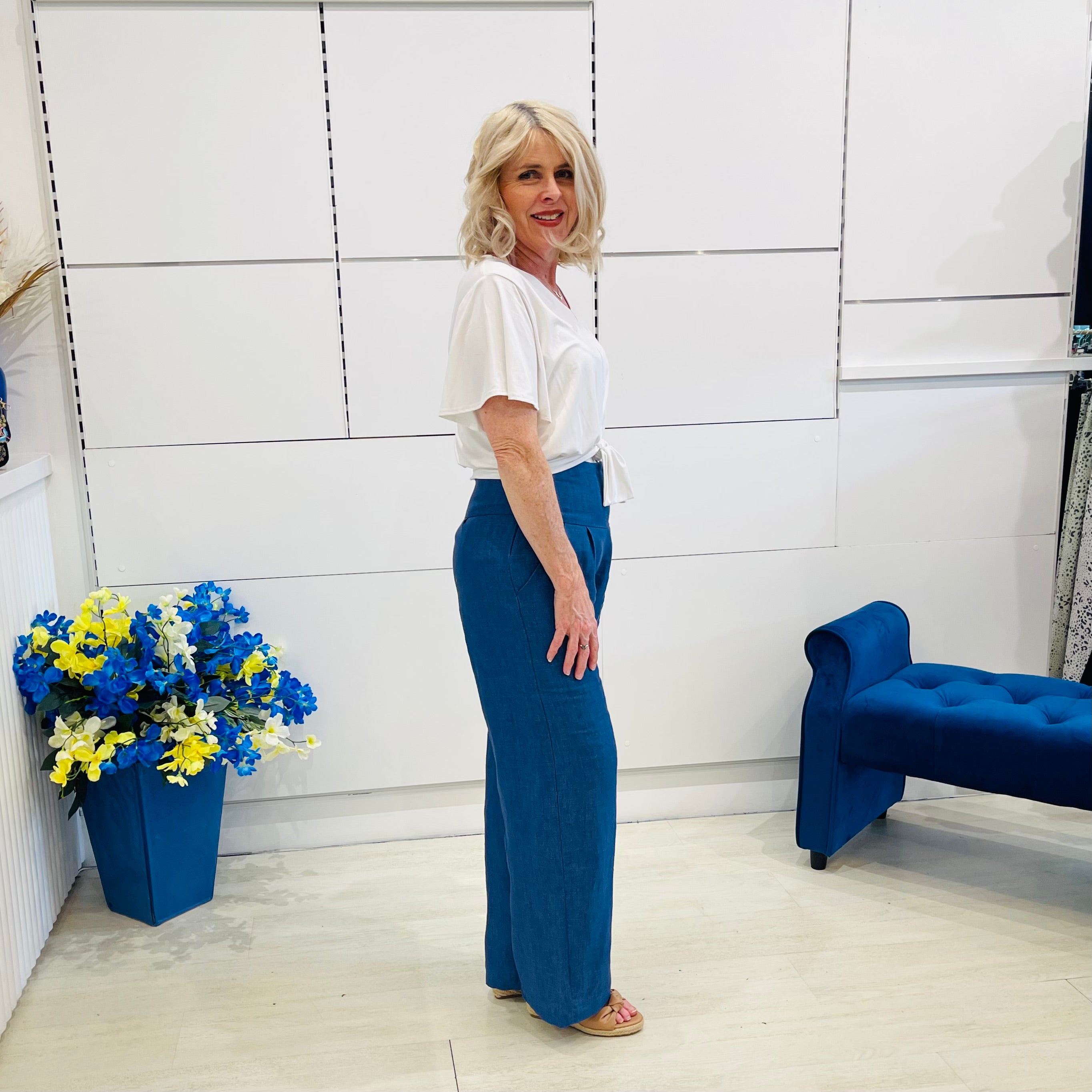 Woman in a white shirt and blue pants standing in a room with a blue bench and flower arrangement.