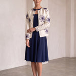 A woman models a cream-colored jacket with purple floral embroidery, worn over a navy blue midi dress, in a minimalist studio setting with beige walls and a concrete floor.