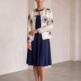 A woman models a cream-colored jacket with purple floral embroidery, worn over a navy blue midi dress, in a minimalist studio setting with beige walls and a concrete floor.