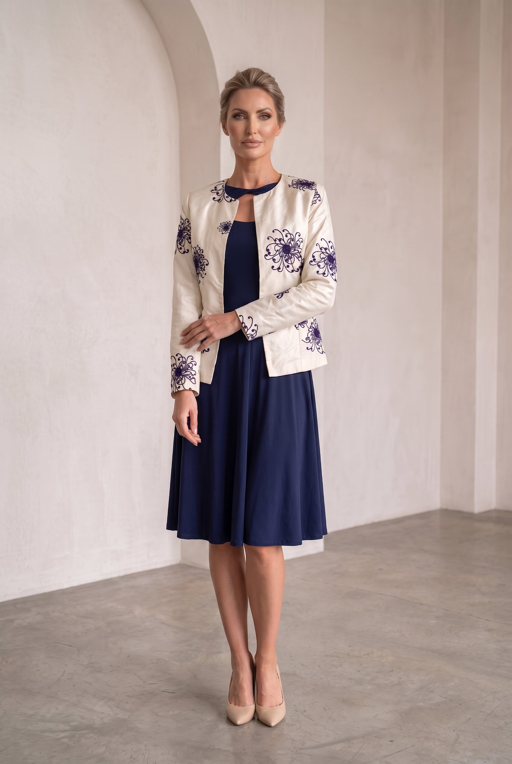 A woman models a cream-colored jacket with purple floral embroidery, worn over a navy blue midi dress, in a minimalist studio setting with beige walls and a concrete floor.