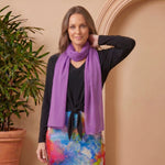 Woman wearing a colorful skirt and purple scarf standing next to a potted plant against a beige wall.