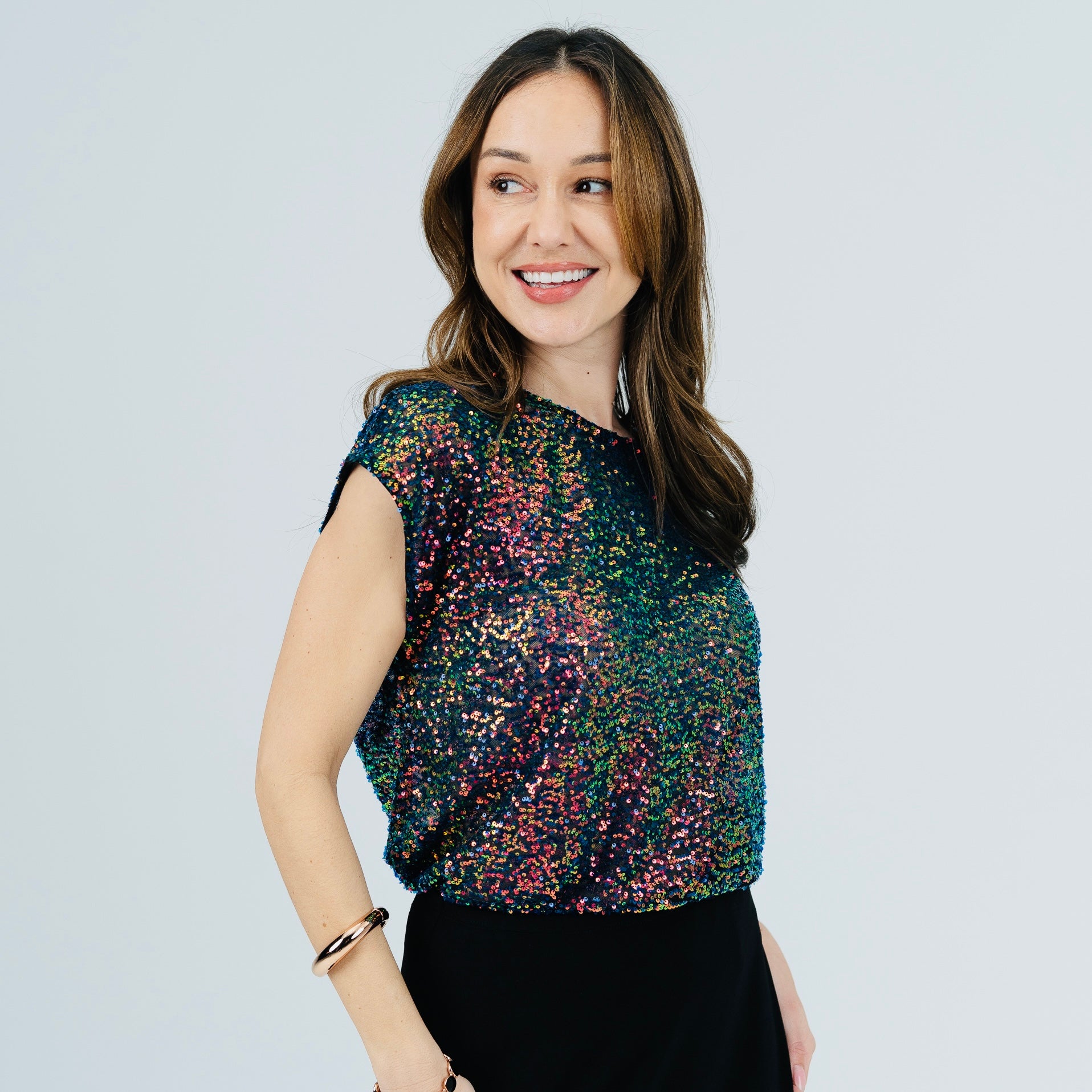 A woman wearing a short-sleeve, crew-neck top covered in multicolor sequins, paired with a black skirt, against a light gray studio background.