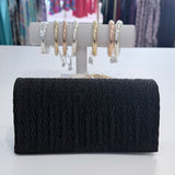 Black clutch bag with textured pattern on a white surface, with jewelry displayed in the background.