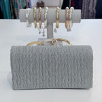 Silver clutch bag with textured pattern and gold handle, displayed in front of a jewelry stand with rings.