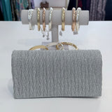 Silver clutch bag with textured pattern and gold handle, displayed in front of a jewelry stand with rings.