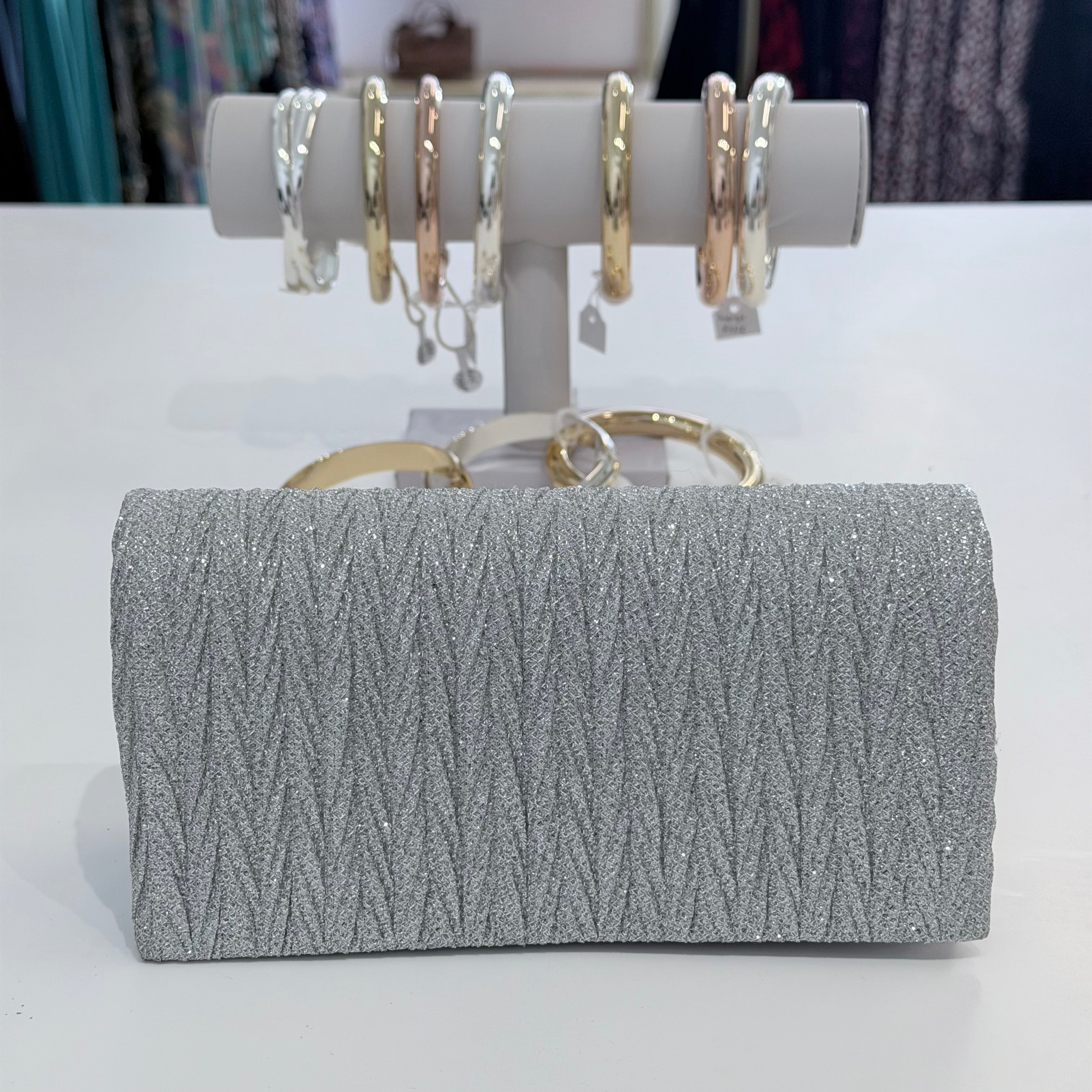Silver clutch bag with textured pattern and gold handle, displayed in front of a jewelry stand with rings.