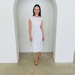 Woman wearing a white dress standing in a minimalistic room with white walls and a concrete floor.