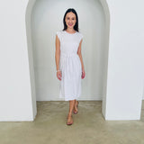 Woman wearing a white dress standing in a minimalistic room with white walls and a concrete floor.