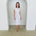 Woman wearing a white dress standing in a minimalistic room with white walls and a concrete floor.