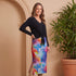 Woman wearing a black top and colorful skirt standing next to a potted plant against a beige wall.