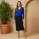Woman wearing a blue top and black skirt standing next to a potted plant against a beige wall.