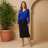 Woman wearing a blue top and black skirt standing next to a potted plant against a beige wall.