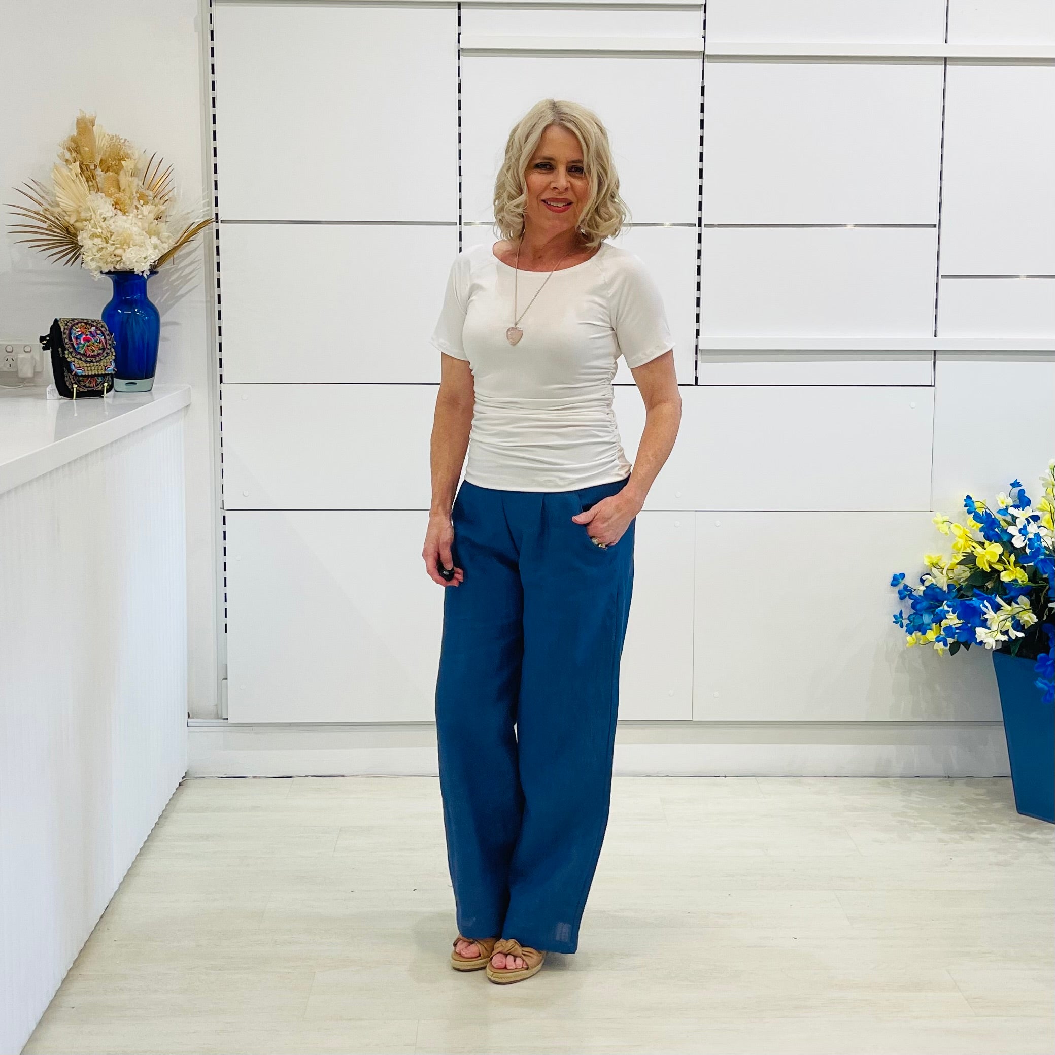 Woman wearing white dressy top, and jeans, standing in a room with white walls and decorative elements.