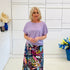 Woman wearing a light purple top and colorful skirt standing in a room with white walls and decorative elements.