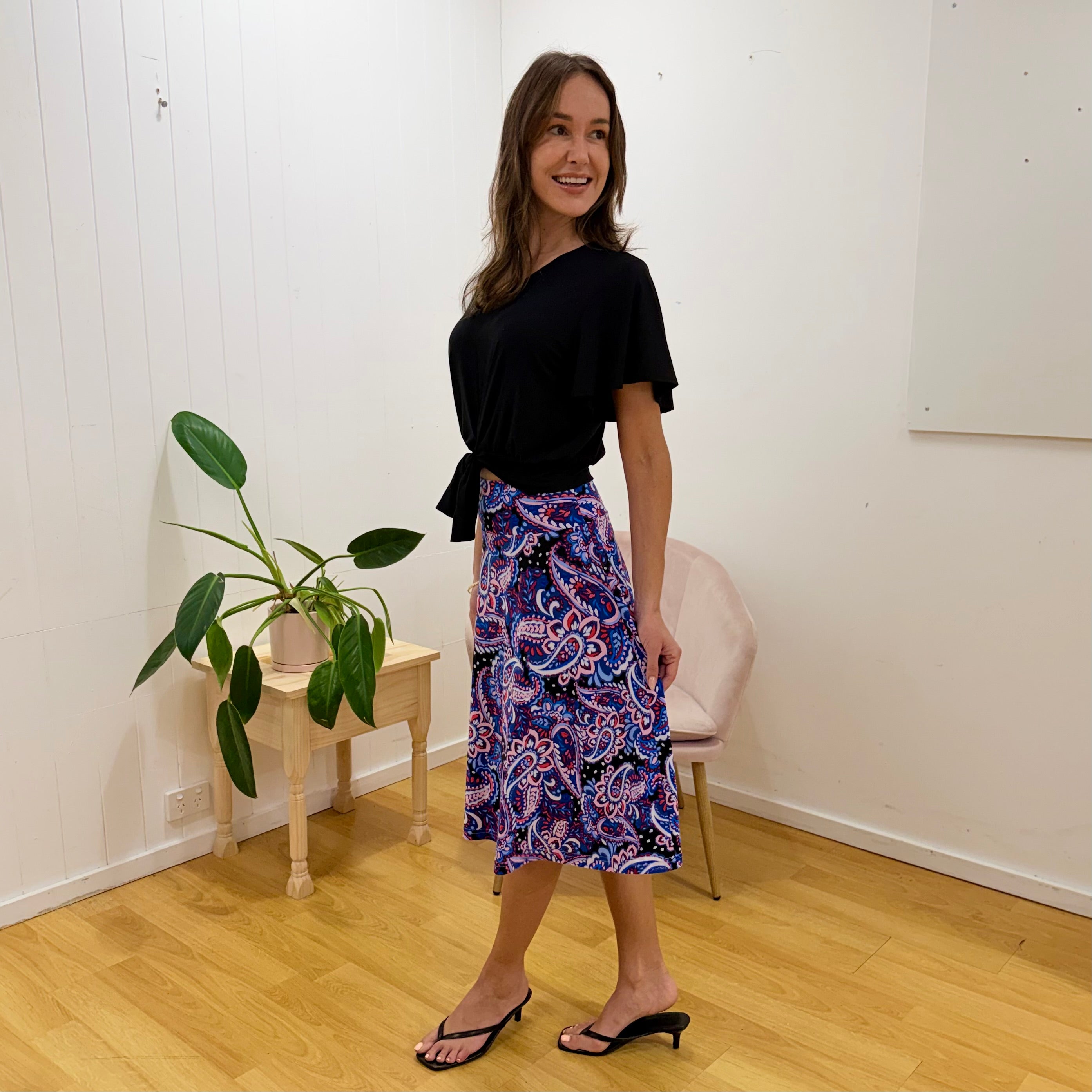 Woman wearing a black top and colorful paisley skirt standing in a room with a plant and chair.