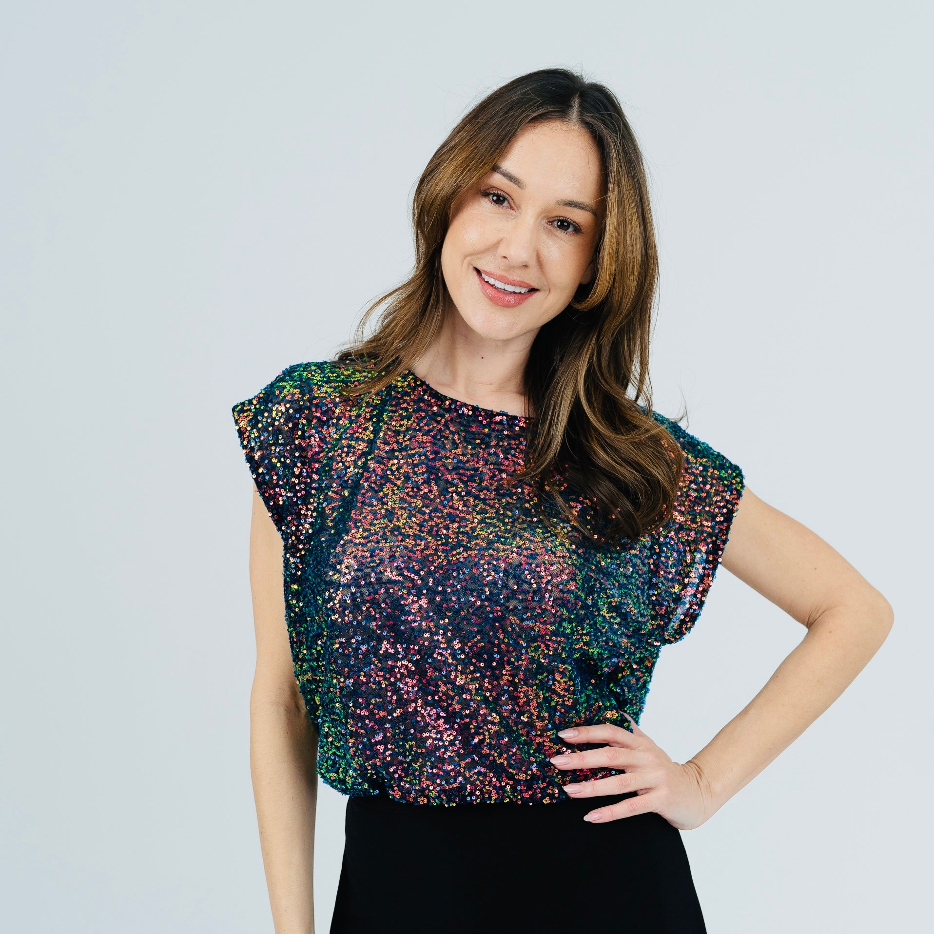 A woman modeling a short-sleeve top covered in multicolor sequins, paired with a black skirt, against a light gray studio background.