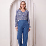 Woman wearing a blue patterned top and blue pants standing in front of a white curtain.