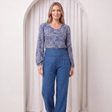 Woman wearing a blue patterned top and blue pants standing in front of a white curtain.