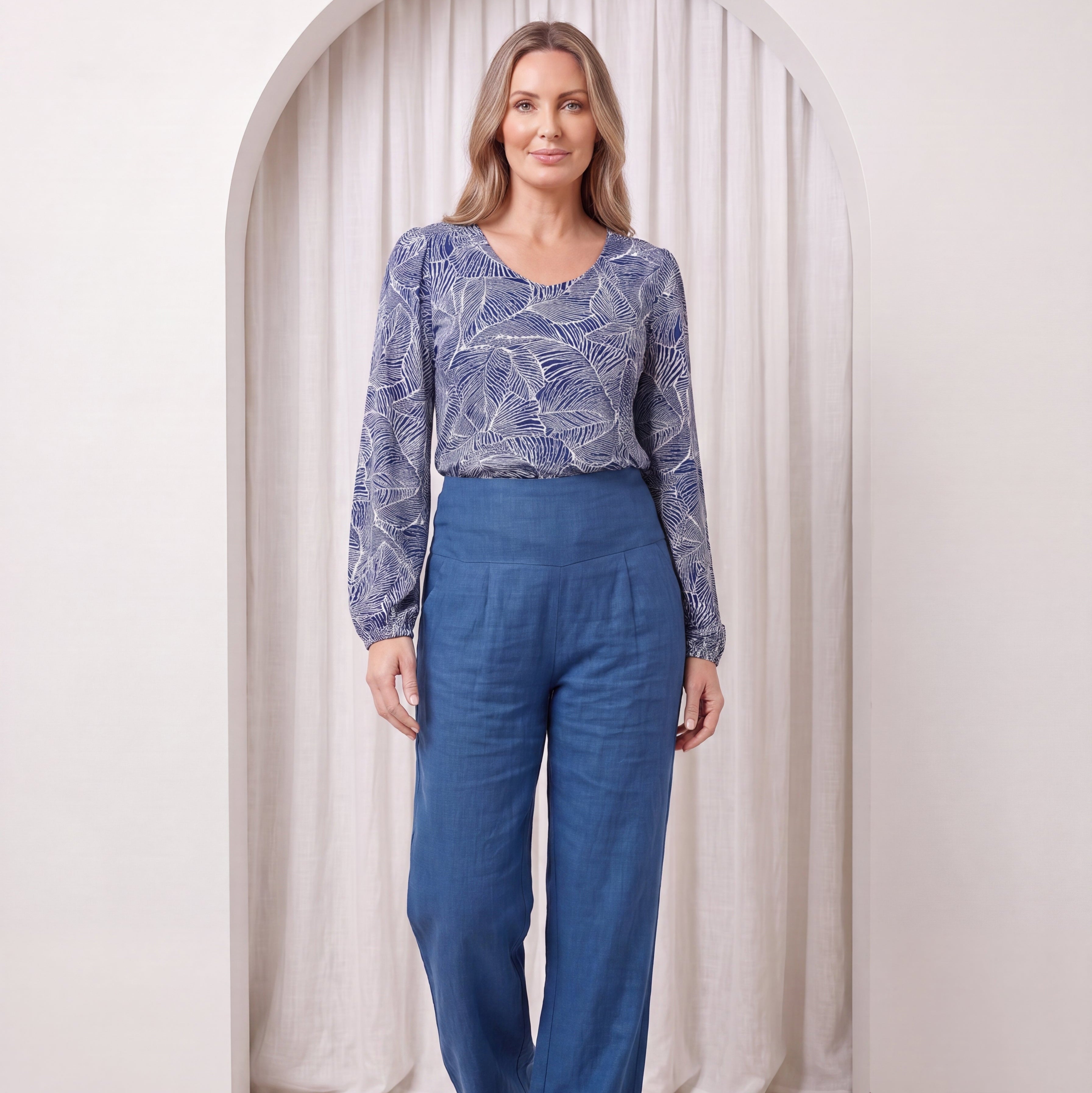 Woman wearing a blue patterned top and blue pants standing in front of a white curtain.