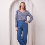 Woman wearing a blue patterned top and blue pants standing in front of a white curtain.