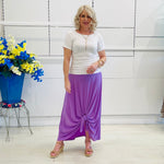 Woman wearing a white top and purple skirt standing in front of a white paneled wall with a blue flower arrangement.