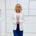 Woman wearing a white blazer over a striped shirt with blue pants against a white wall.