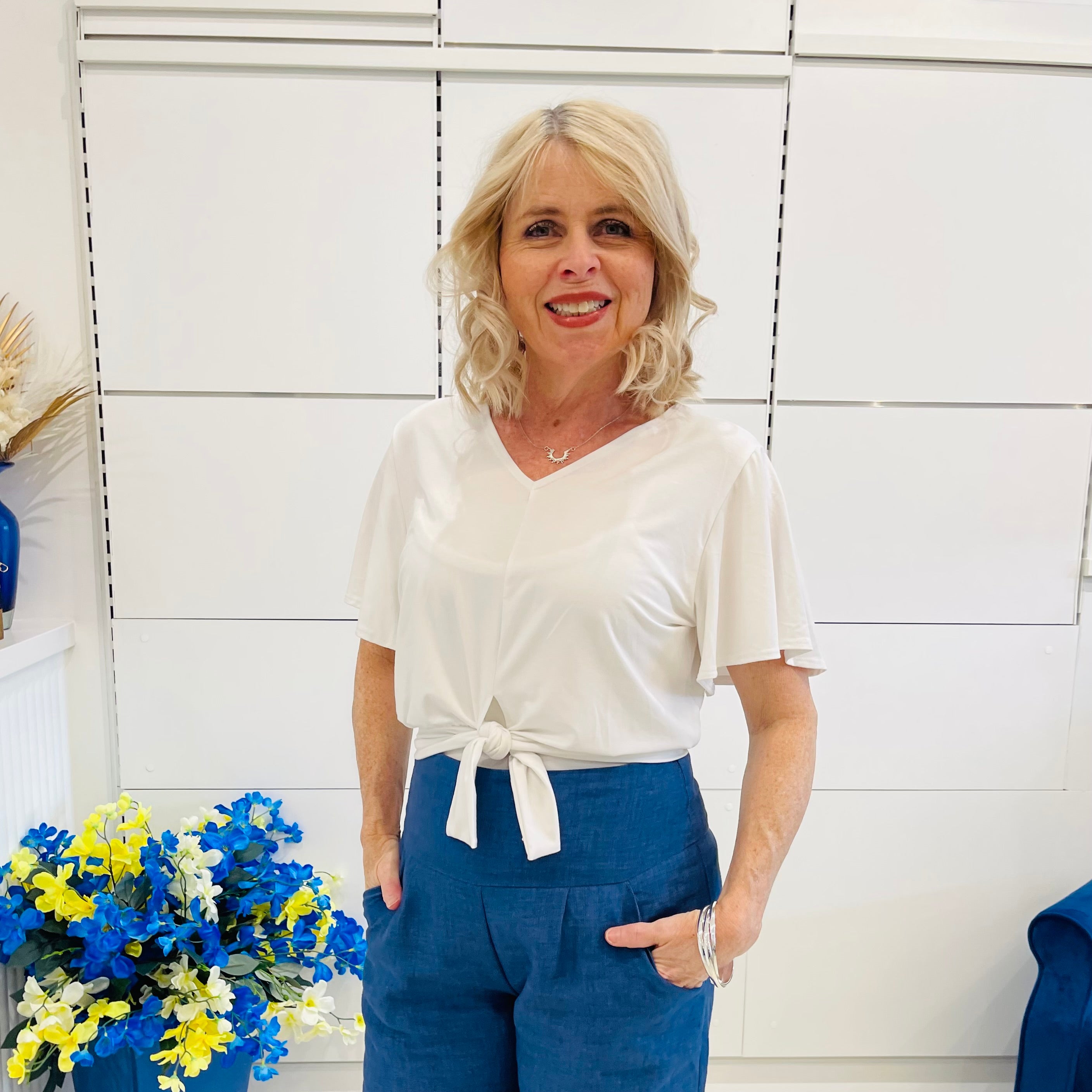 Woman wearing a white top and blue pants standing in front of a white tiled wall with flowers to her left.