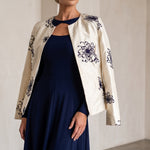 A woman models a navy blue midi dress with a keyhole cutout, styled with an off-white embroidered jacket featuring dark blue floral patterns, in a minimalist beige-walled room.