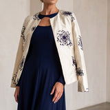 A woman models a navy blue midi dress with a keyhole cutout, styled with an off-white embroidered jacket featuring dark blue floral patterns, in a minimalist beige-walled room.