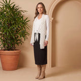 Woman in a white cardigan and black skirt standing next to a potted plant against a beige wall.