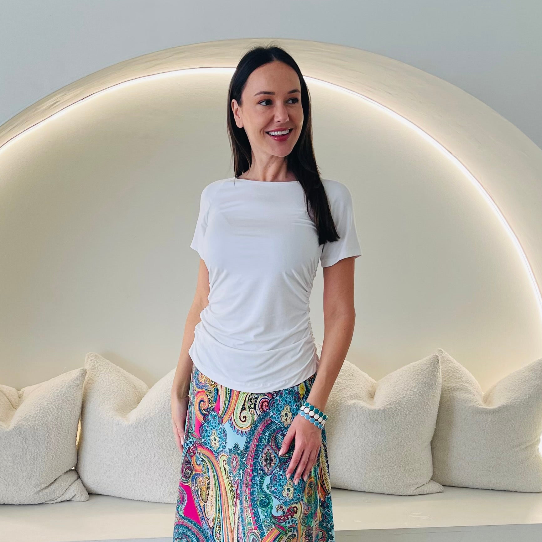 Woman wearing a white top and colorful skirt standing in front of a modern, curved wall.