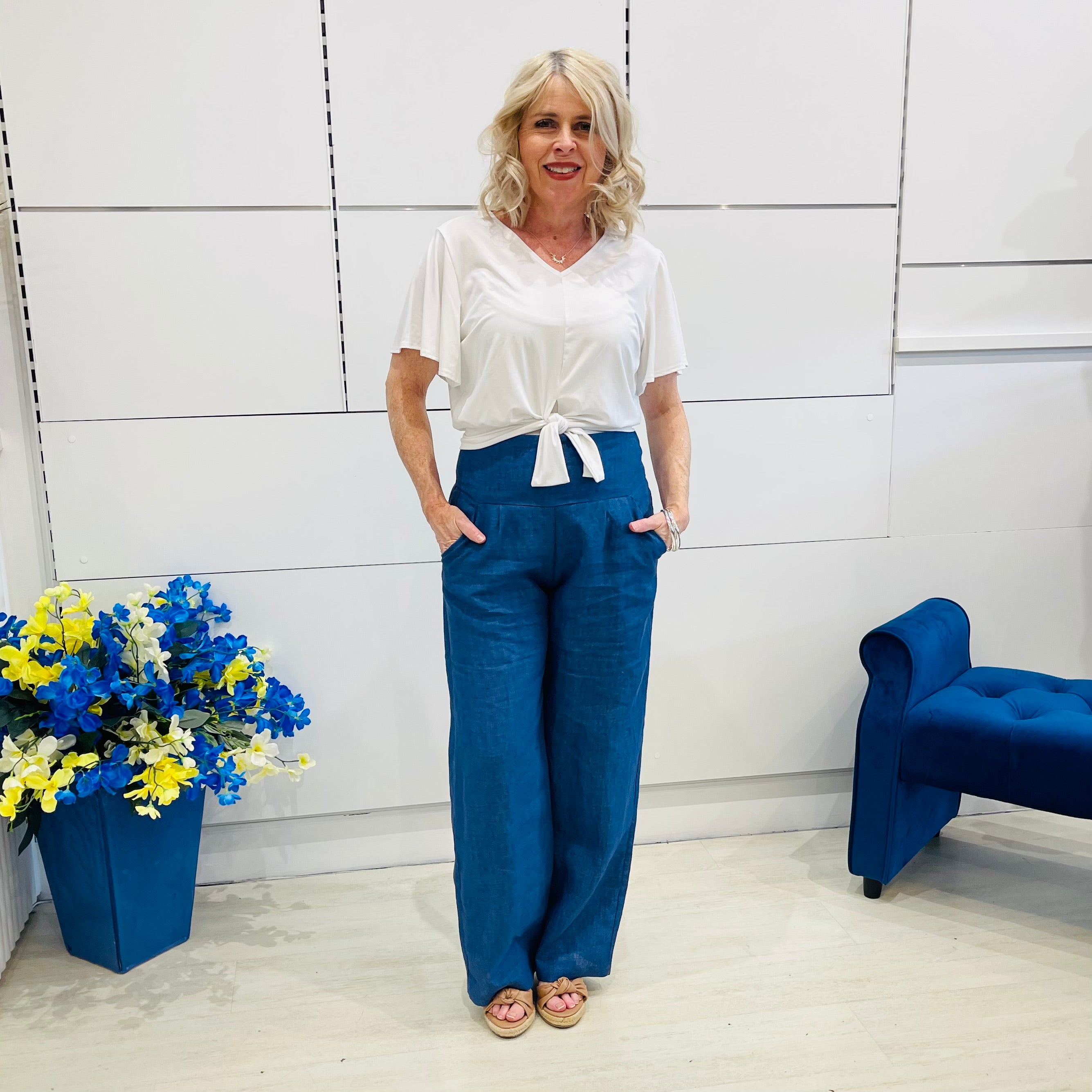 Woman wearing a white top and blue pants standing in a room with a blue chair and flower arrangement.