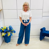 Woman in a white top and blue pants standing in a room with a blue chair and flower arrangement.