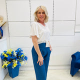 Woman in a white top and blue pants standing next to a blue chair and flower arrangement.