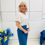 Woman in a white blouse and blue pants standing in a room with a blue chair and floral arrangement.