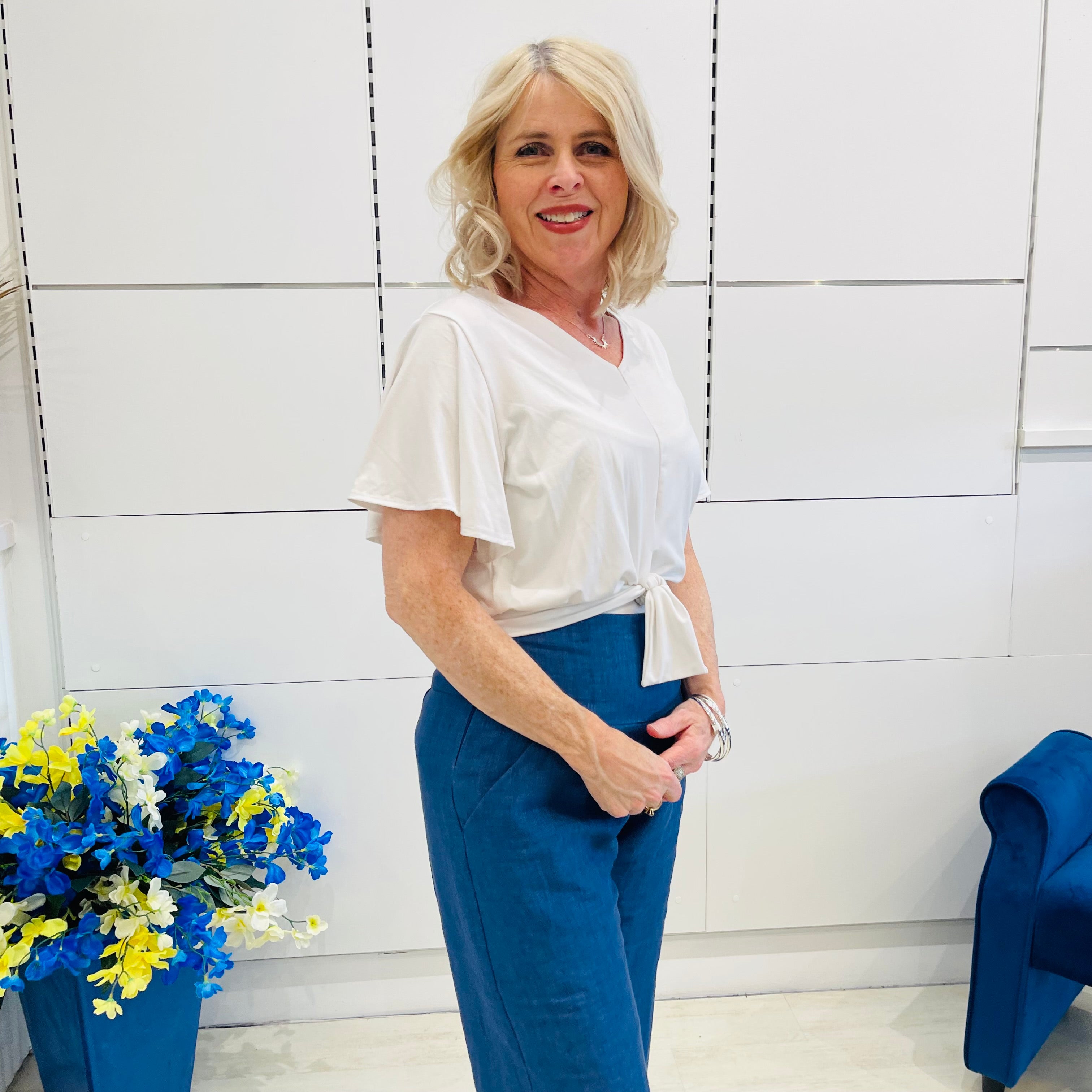 Woman in a white blouse and blue pants standing in a room with a blue chair and floral arrangement.
