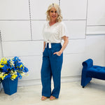 Woman in a white top and blue pants standing in a room with a blue chair and flower arrangement.