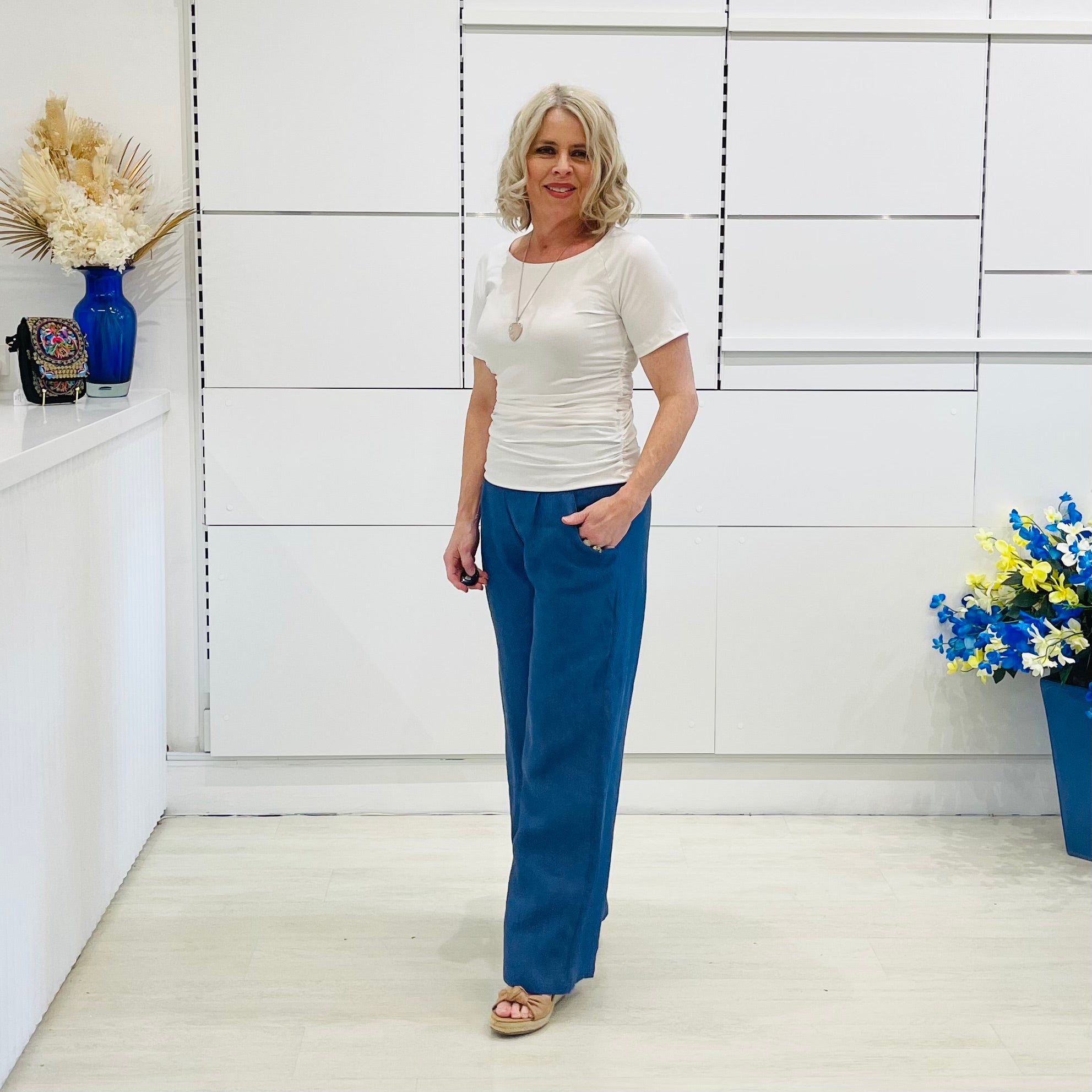 Woman wearing a white top and blue pants standing in a room with white walls and a shelf.