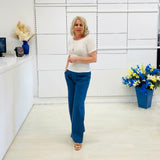 Woman in a white top and blue pants standing in a room with white walls and a vase of flowers.